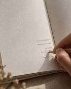 A close-up of a hand holding a wooden pencil and writing on the inside cover of a textured off-white notebook or journal. The cover features printed text including 'Written by Vinda Dwived' and 'Illustrated by Urja Dwived'.